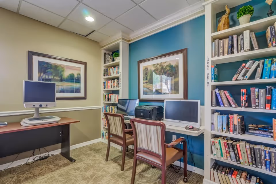 Small senior living community computer/library room with two desktop computers at a wall desk, bookshelves filled with books, framed artwork, and chairs.