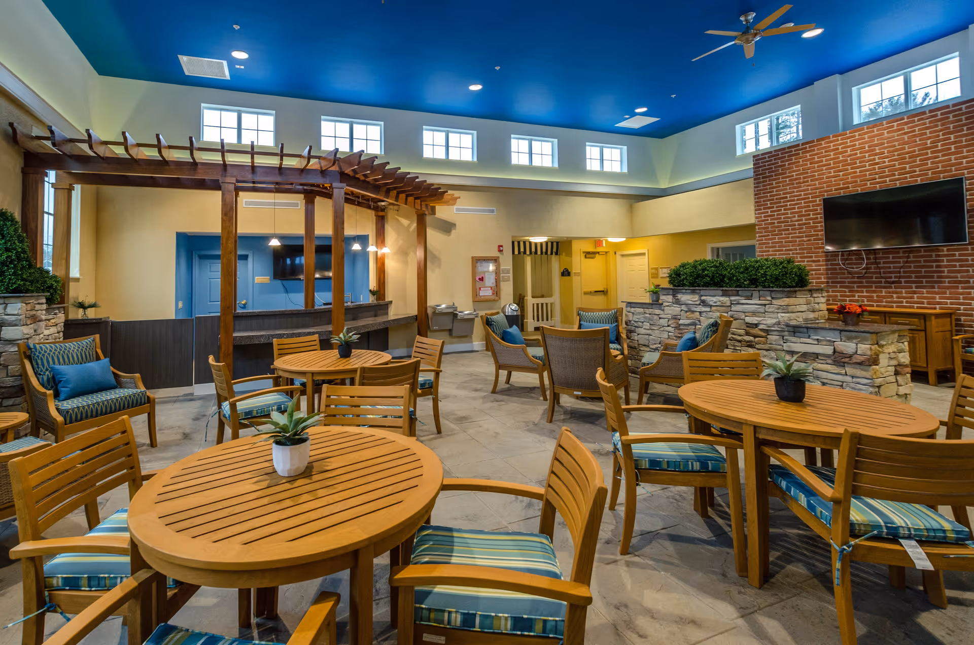 A spacious senior living common area with wooden round tables and chairs featuring blue striped cushions. There are also cushioned armchairs with blue pillows arranged near a stone partition with greenery. The room has a blue ceiling with recessed lighting and ceiling fans, multiple windows near the ceiling for natural light, and a large flat-screen TV mounted on a brick wall.