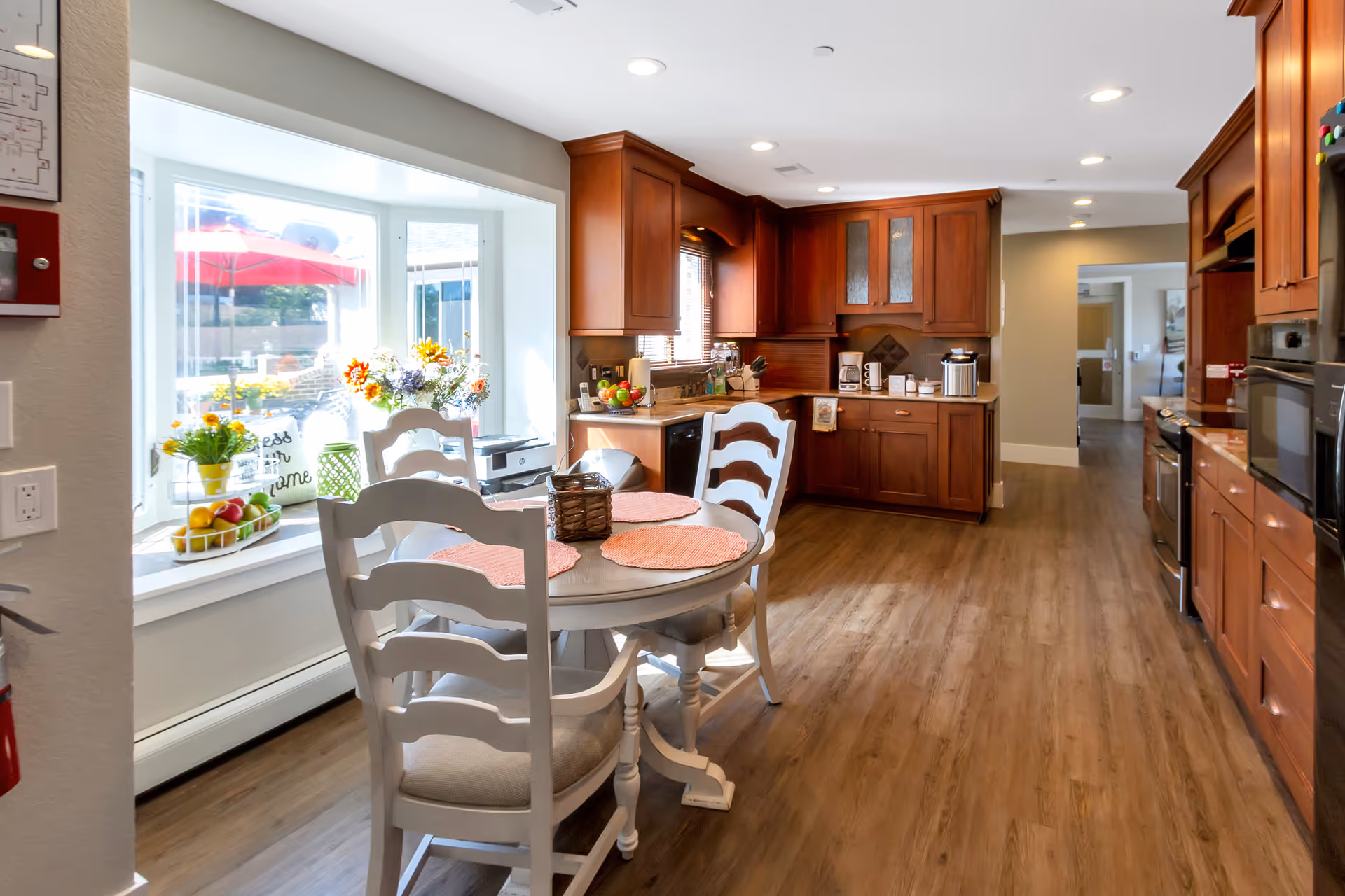 A bright kitchen area with wooden cabinets and a round dining table with four white chairs. The table has four orange placemats and a small decorative basket. A large window with a seating ledge displays flowers, fruit, and decorative items. The kitchen has modern appliances and wood flooring, with recessed lighting in the ceiling.