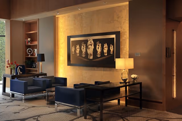 A modern living room area with dark blue armchairs, a black coffee table, and a black console table with a lamp and flowers. The wall behind features a large framed artwork with multiple white sculptures on a black background. The room has warm lighting and a patterned carpet.