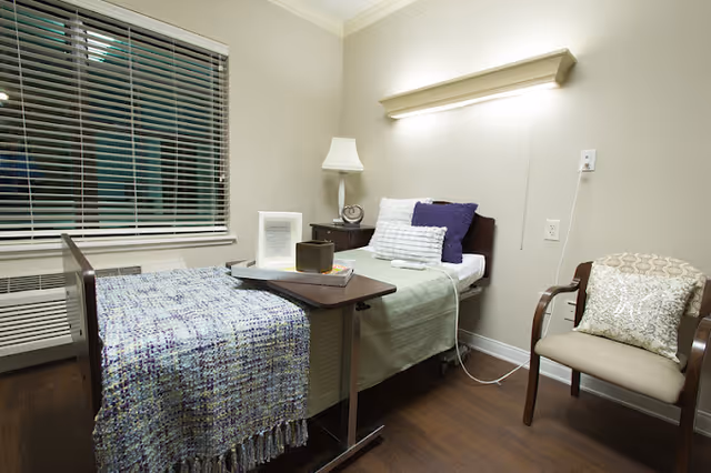 A small, neatly arranged bedroom in a senior living facility with a single bed covered with a green blanket and a blue patterned throw. The bed has white and purple pillows and a tray table with items on it. There is a wooden chair with a decorative pillow, a nightstand with a lamp and a clock, and a window with blinds. The room has wooden flooring and light-colored walls.