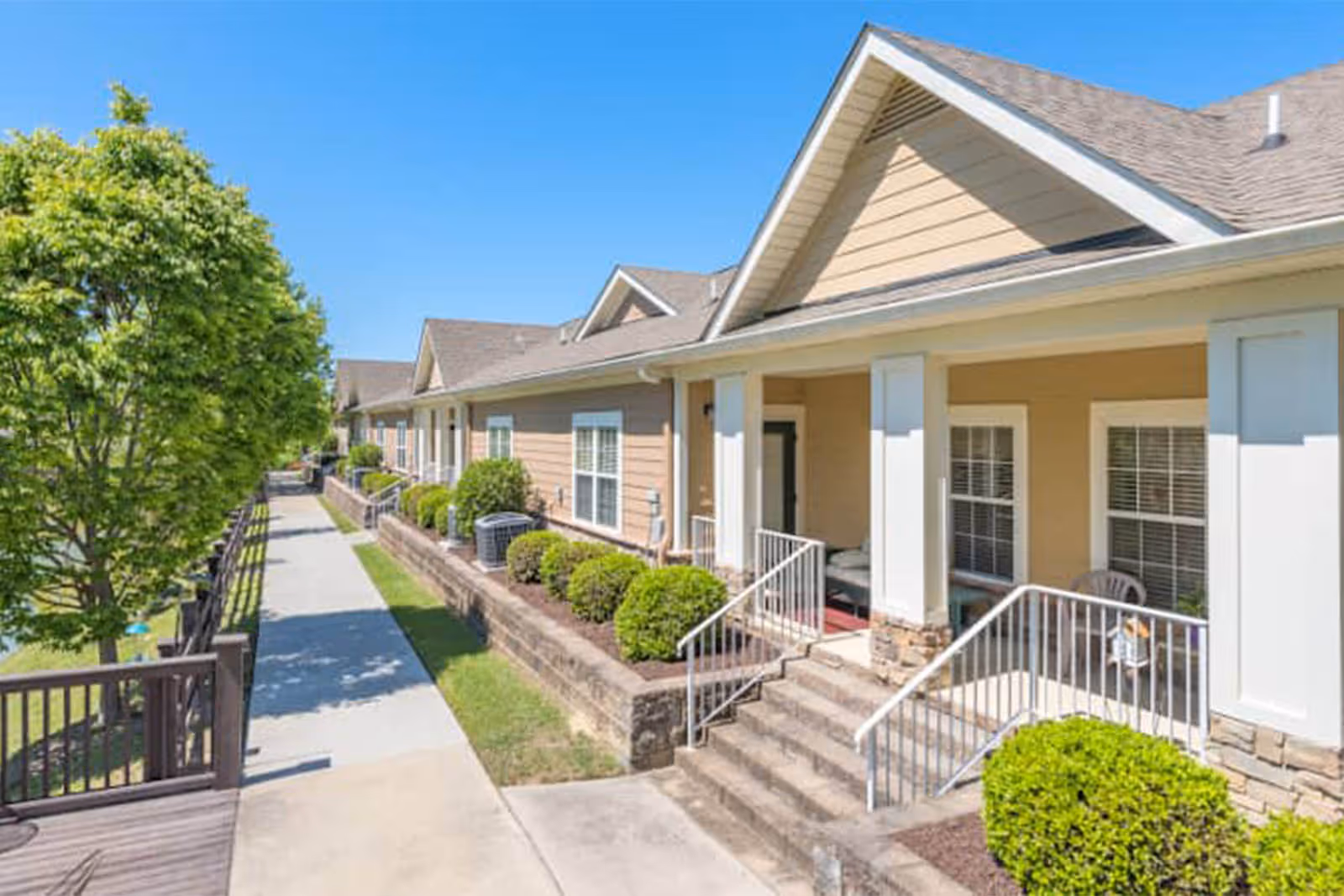 Exterior view of a single-story residential building with beige siding and white trim, featuring a covered porch with steps and railings. There are neatly trimmed bushes along the building and a paved walkway beside it, with a green tree on the left under a clear blue sky.