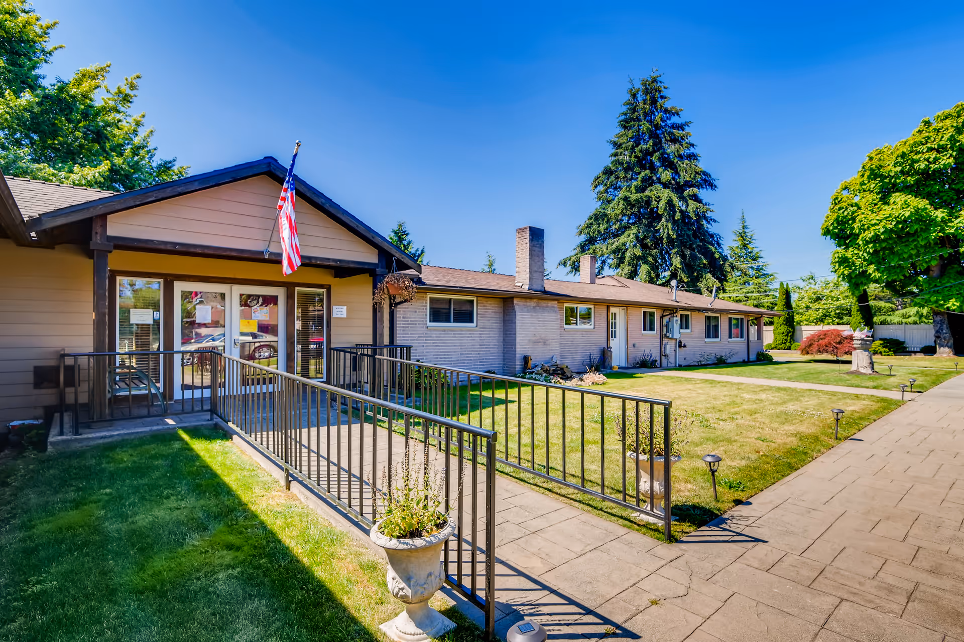 Exterior view of a single-story senior living facility building with a ramp leading to the entrance. The building has beige siding and brick walls, with an American flag mounted above the entrance. The surrounding area includes a well-maintained lawn, trees, and a paved walkway under a clear blue sky.