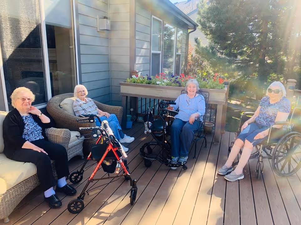 Four elderly women sitting outside on a wooden deck. Two women are seated on cushioned wicker chairs, one woman is sitting on a metal chair, and one woman is in a wheelchair. There are walkers in front of two of the women. The deck has a planter box with colorful flowers and the background shows part of a house and trees with sunlight streaming through.