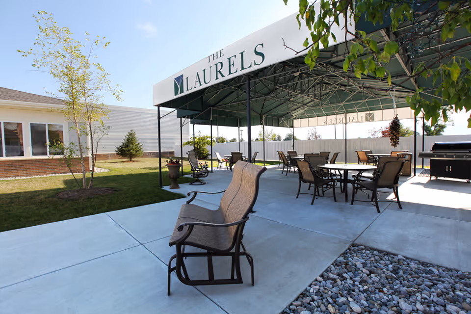 Outdoor patio area at The Laurels with a covered seating area featuring multiple tables and chairs, a grill, and surrounding greenery including trees and grass under a clear blue sky.