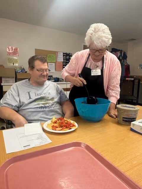 A woman wearing a pink cardigan and black apron is serving food from a large blue bowl to a man sitting at a table. The man is wearing glasses and a gray t-shirt with text on it. On the table, there is a plate with food, a container of Country Crock spread, some papers, and a red tray. The setting appears to be a communal dining or activity area within a facility.