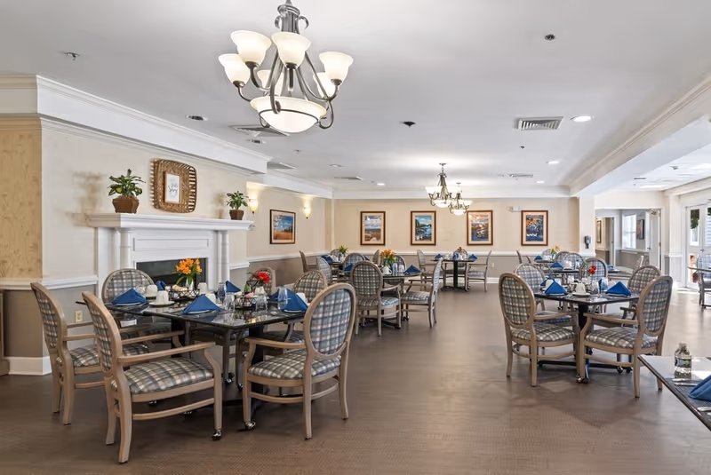 A spacious dining room with multiple round tables set with blue napkins, glassware, and small flower arrangements. The room features plaid cushioned chairs, a white fireplace with plants and framed art above it, beige walls with framed pictures, and elegant chandeliers hanging from the ceiling.