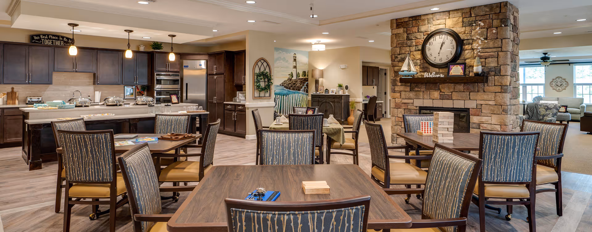 A spacious and well-lit common area in a senior living facility featuring multiple wooden tables and chairs arranged for social activities. The room includes a kitchen area with dark wood cabinets, stainless steel appliances, and pendant lighting. A stone fireplace with a large clock and decorative items is visible, along with a mural of a lighthouse on one wall. The space has a warm and inviting atmosphere with neutral tones and comfortable seating.