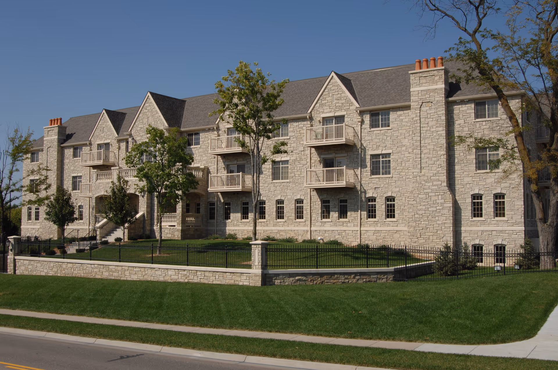 Large stone-faced three-story residential building with balconies, landscaped lawn, and iron fence viewed from the street.