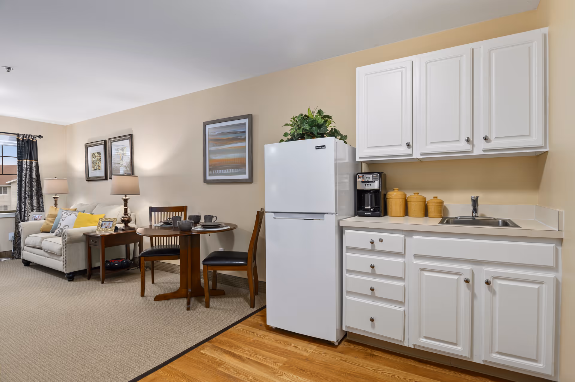 Interior view of a senior living facility apartment showing a small kitchen area with white cabinets, a white refrigerator, a coffee maker, and three yellow canisters on the countertop. Adjacent to the kitchen is a dining area with a round wooden table set with dishes and two chairs. In the background, there is a living area with a beige sofa, side table with a lamp, framed artwork on the wall, and a window with curtains.
