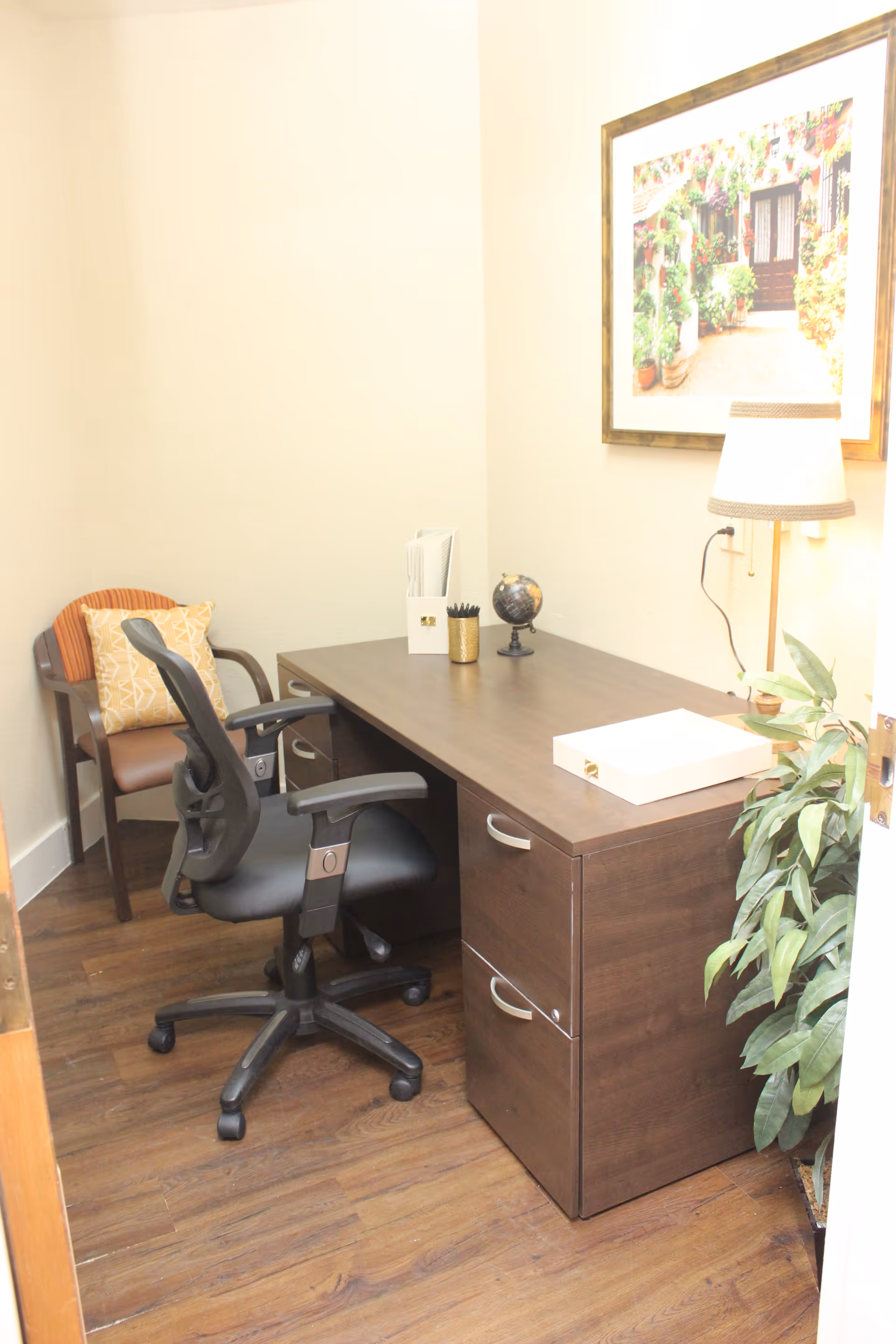A small office space with a dark wooden desk, a black ergonomic office chair, and a wooden chair with a cushion. On the desk are a small globe, a white file holder with papers, a gold pencil holder with pens, a white box, and a table lamp. A framed picture of a garden scene hangs on the wall, and a green plant is placed beside the desk on the wooden floor.