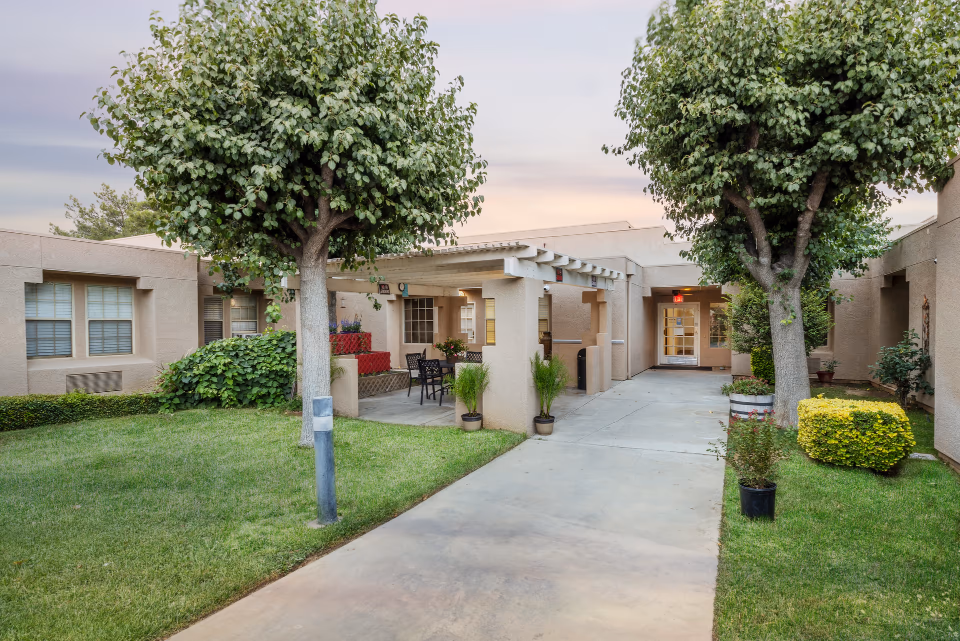 Outdoor courtyard and entrance of a single-story senior living facility with a paved walkway, pergola seating area, trees, and manicured lawns.