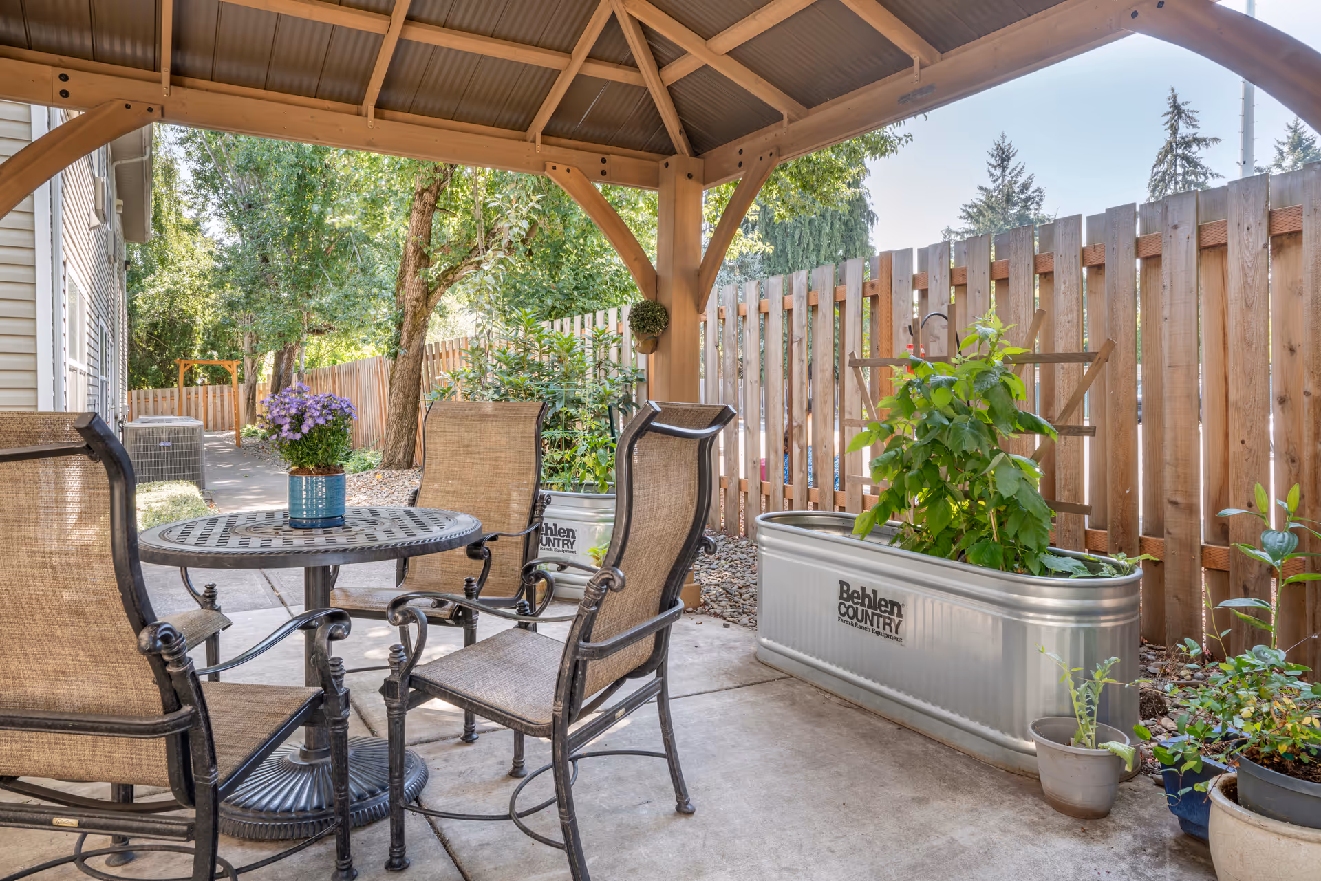 Covered outdoor patio area with a round metal table and four cushioned chairs. There are several potted plants and a large metal planter with green plants along a wooden fence. Trees and greenery are visible beyond the fence under a clear sky.