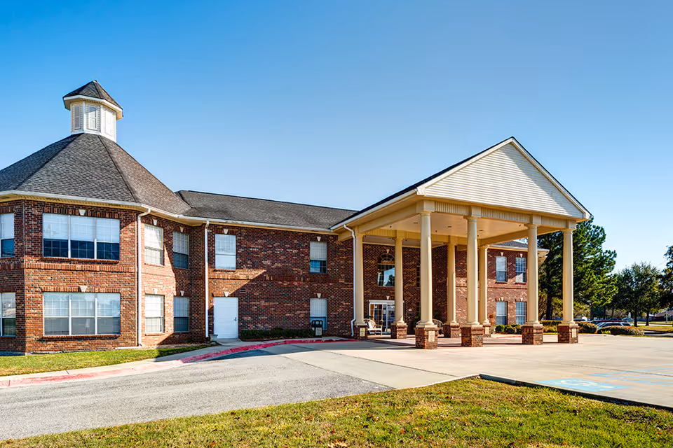 Brick two-story senior living building front with a columned covered entrance and cupola under a clear blue sky.