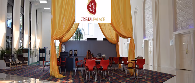 Interior view of a spacious room with high ceilings and large windows. The room features a seating area with red chairs and tables under a yellow draped canopy. There are additional chairs along the walls and plants near the windows. The floor is covered with a colorful patterned carpet, and the walls are white with decorative molding.