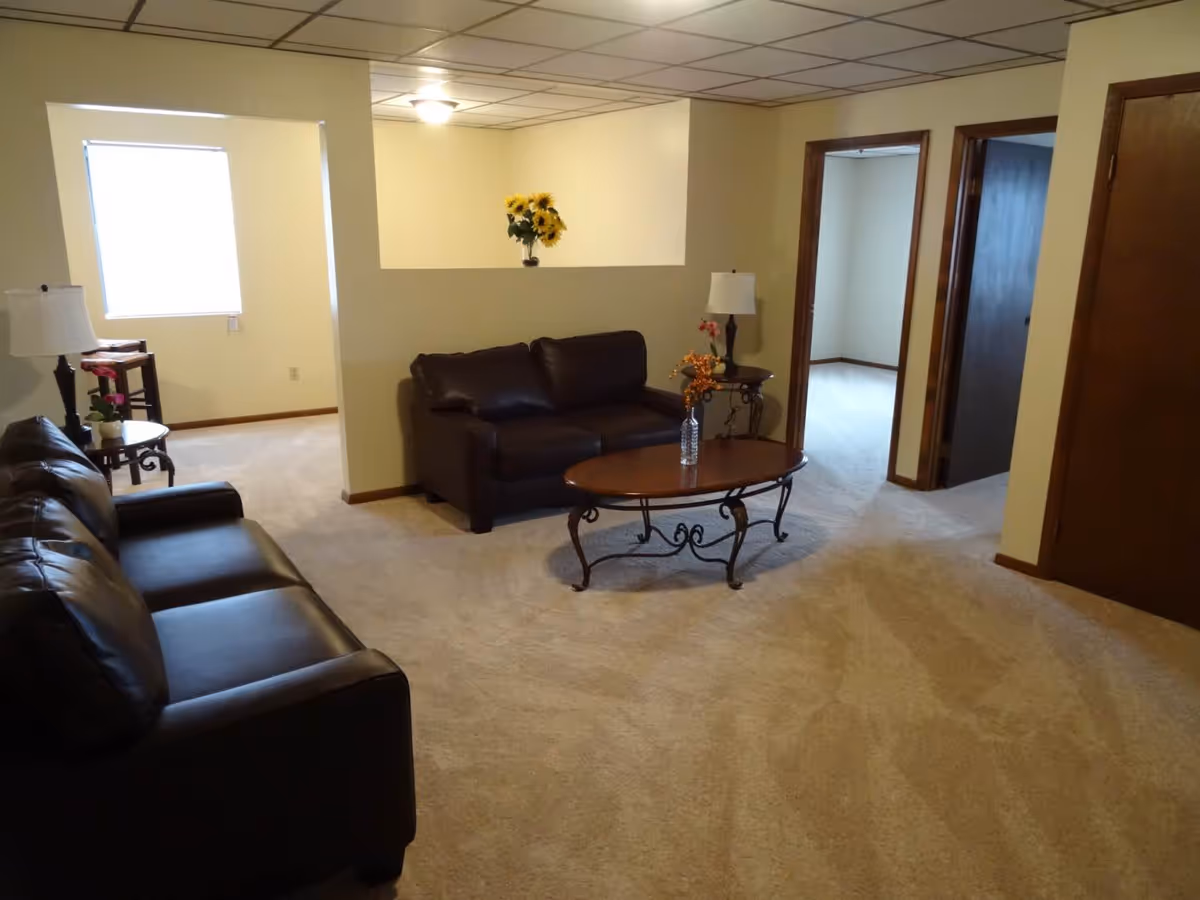 A living room with beige carpet and cream-colored walls featuring two dark brown leather sofas, a wooden coffee table with a vase of flowers, two side tables with lamps, and a window letting in natural light. There are two open doorways leading to other rooms.