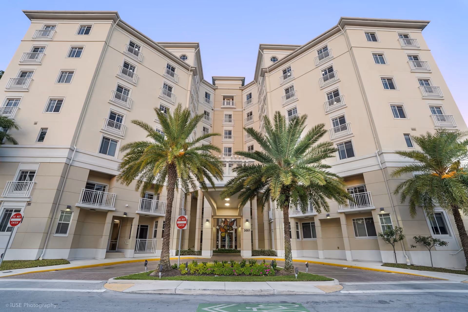 Exterior view of a multi-story senior living facility building with beige walls and multiple windows. The entrance is framed by two tall palm trees and a landscaped area with flowers. There are 'Do Not Enter' signs on either side of the driveway leading to the entrance.
