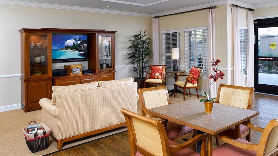 A cozy living room area in a senior living facility featuring a beige sofa facing a wooden entertainment center with a TV displaying a beach scene. There is a square table with four wooden chairs in the foreground, a potted orchid on the table, and two armchairs with red floral cushions near a window with white blinds. A tall green plant and a floor lamp are also visible, along with a glass door leading outside.