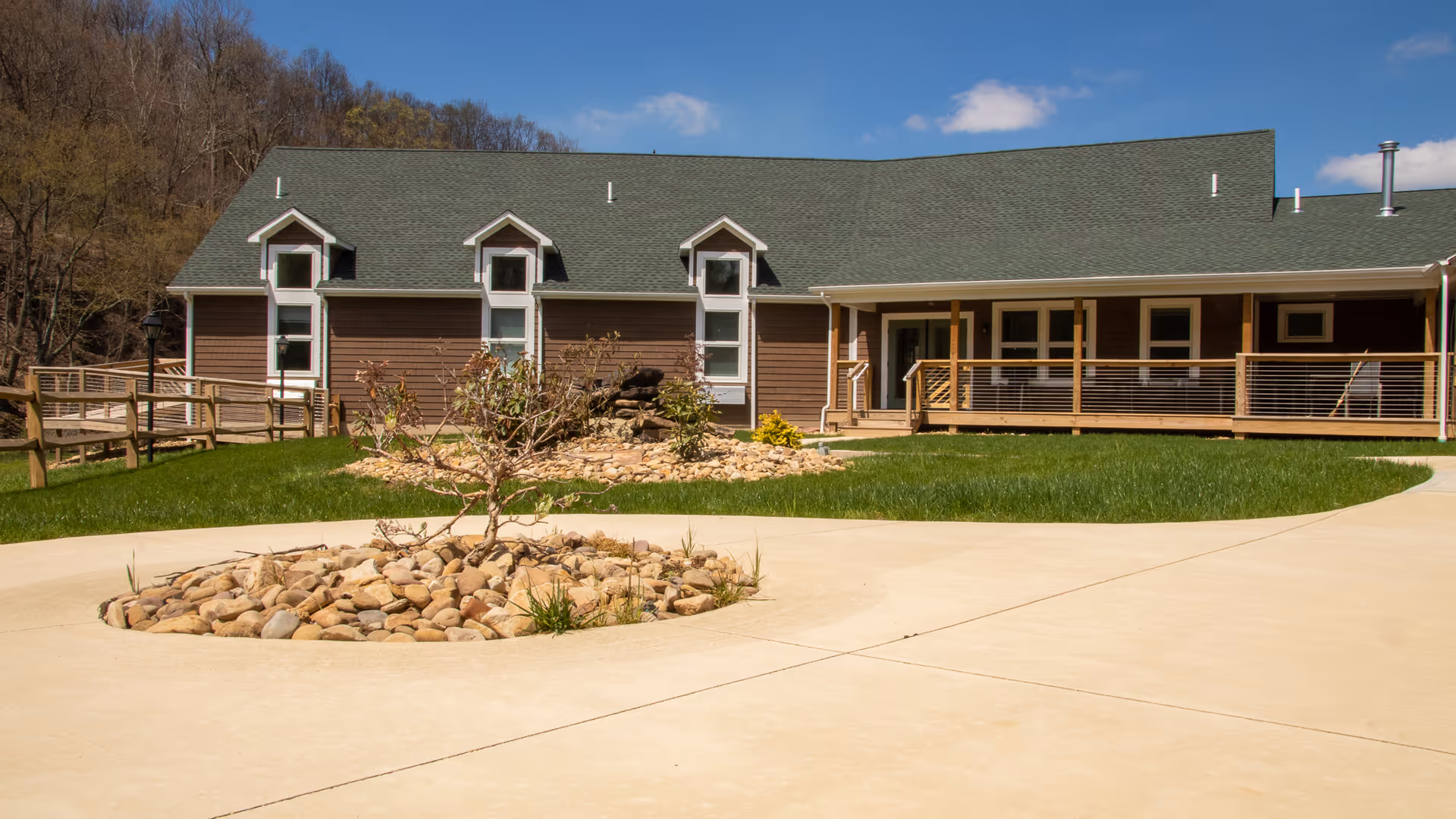 Exterior view of a single-story brown building with a green roof, surrounded by a concrete pathway and landscaped areas with rocks and small plants. The building has multiple windows and a covered porch with wooden railings. Trees and a blue sky with some clouds are visible in the background.