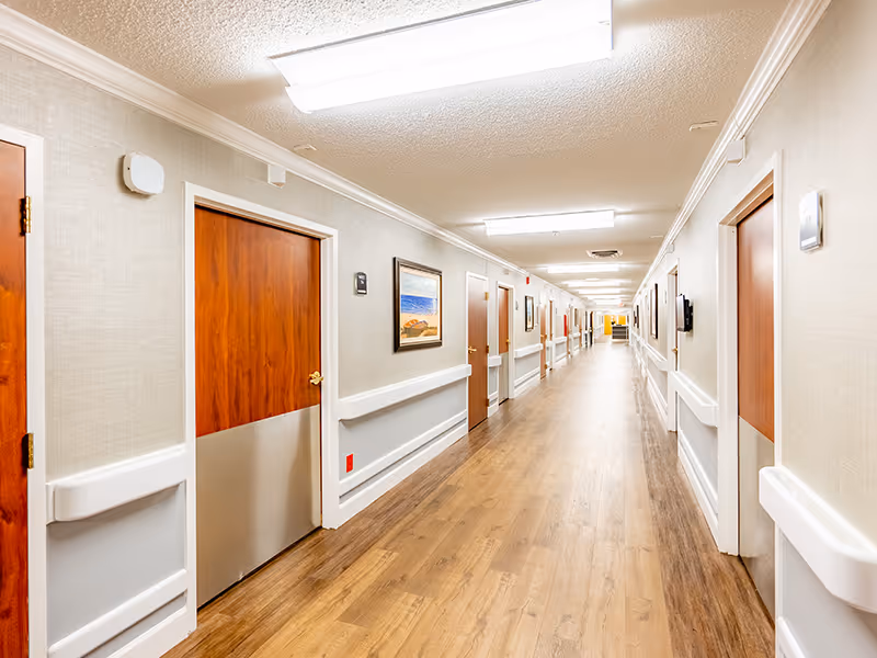 A well-lit hallway in Brushy Creek Post Acute, featuring wooden doors on either side, framed artwork on the walls, and a smooth wooden floor.