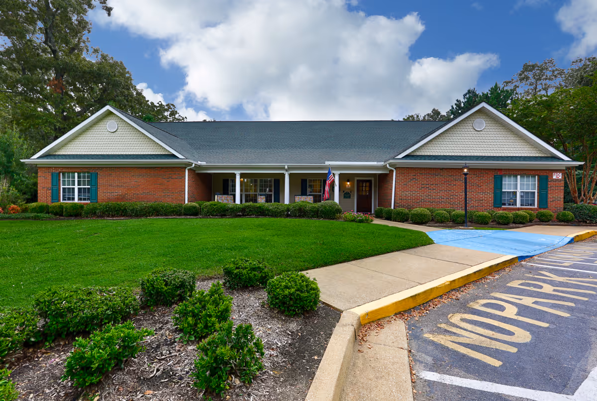Front exterior view of a single-story brick building with green shutters and a gray roof, surrounded by a well-maintained lawn and bushes under a partly cloudy sky. A sidewalk leads to the entrance with an American flag displayed near the door. A parking area with a 'NO PARKING' zone is visible in the foreground.