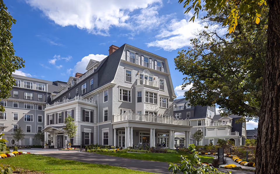Large elegant multi-story senior living building with a covered entrance, white siding, balconies, and landscaped grounds under a blue sky.
