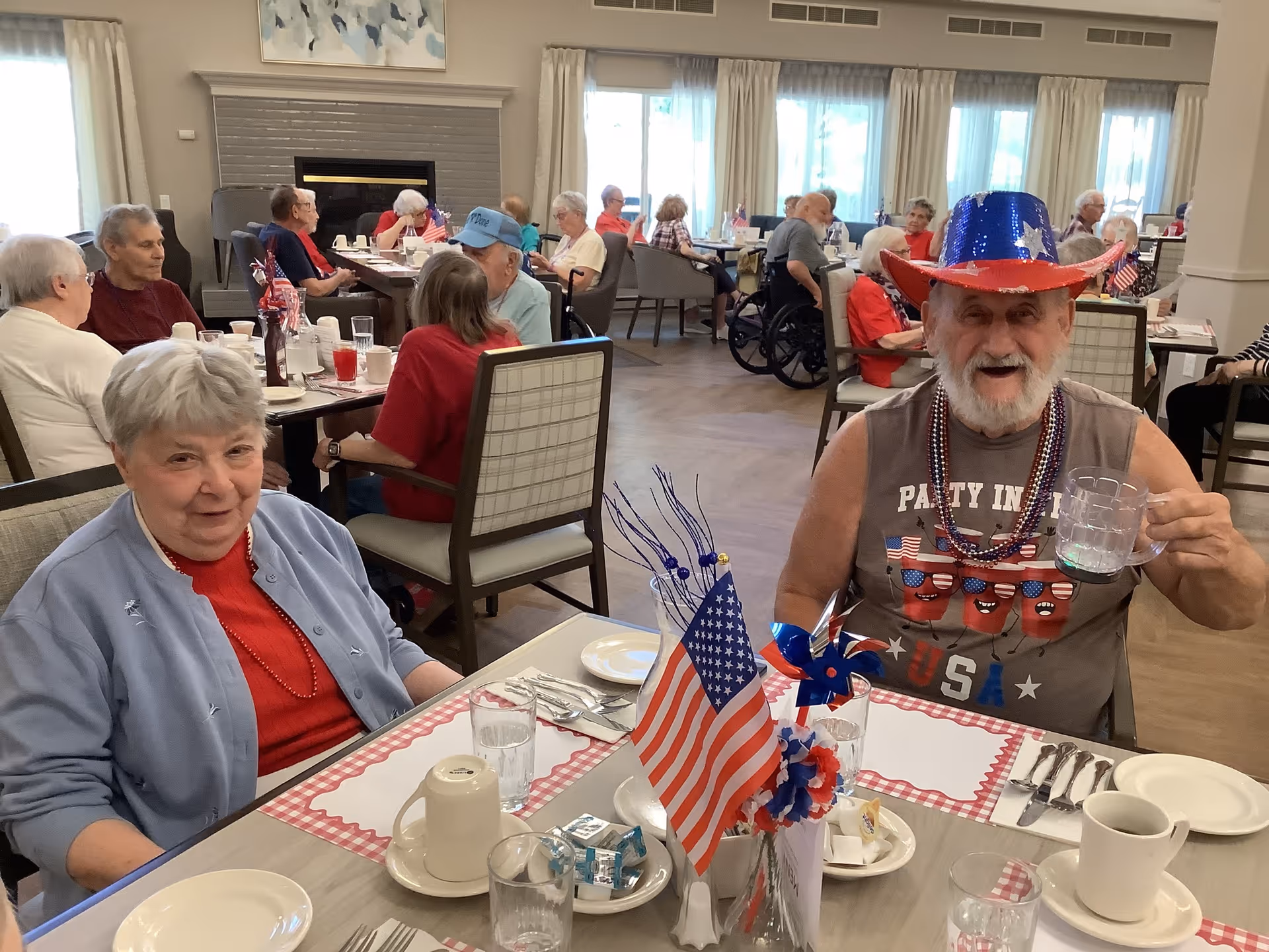 A group of elderly people sitting at tables in a dining room decorated with patriotic American flags and red, white, and blue decorations. In the foreground, a smiling elderly man wearing a blue and red star-spangled hat and a sleeveless shirt with USA graphics holds up a clear mug. Next to him, an elderly woman in a light blue cardigan and red shirt sits at the table. Other seniors are seated and engaged in conversation in the background.