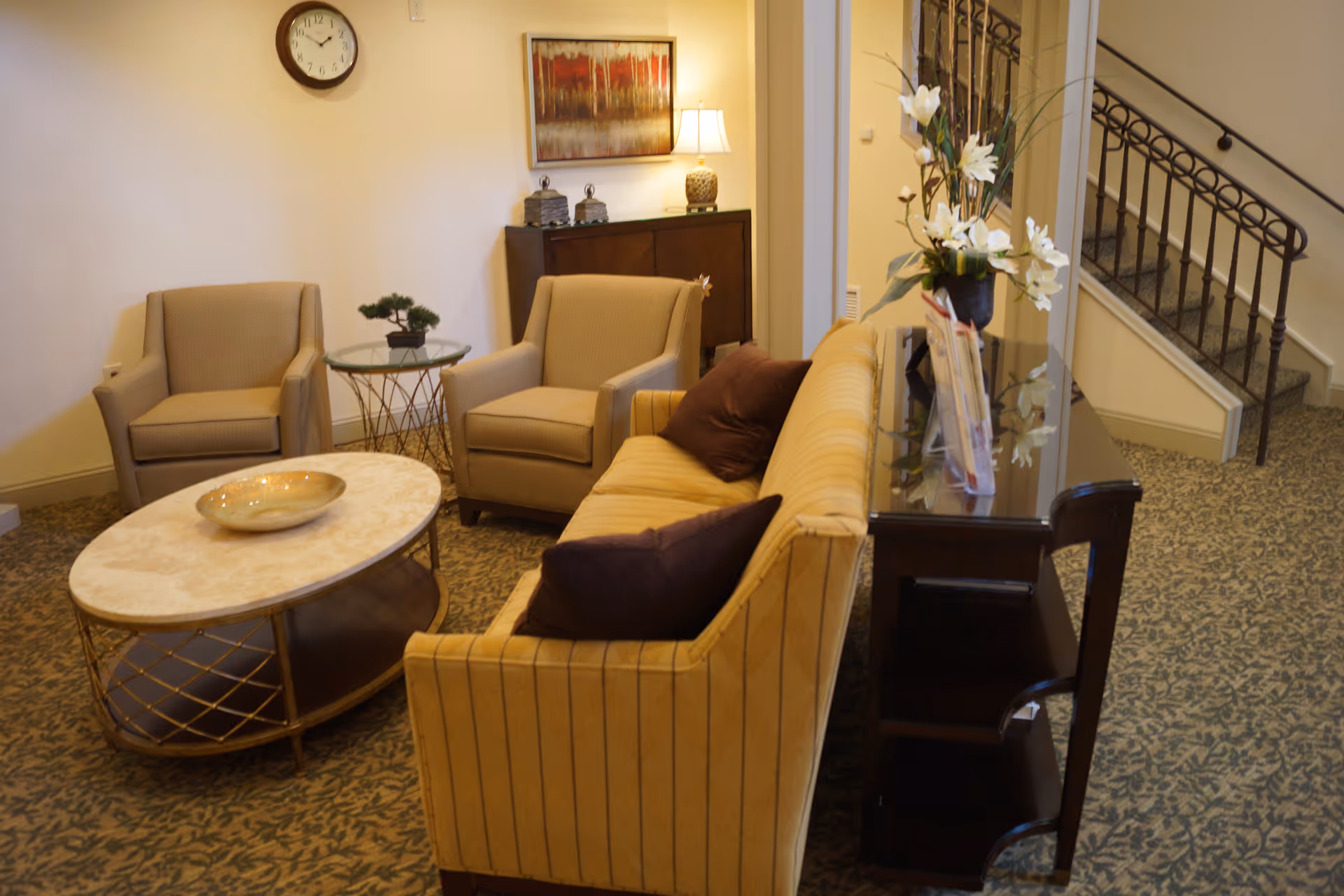 A cozy living room area with a yellow striped sofa, two beige armchairs, a round marble coffee table with a decorative bowl, a small glass side table with a plant, a dark wooden console table with a vase of white flowers, a wall clock, a framed painting, and a staircase with a decorative railing in the background.