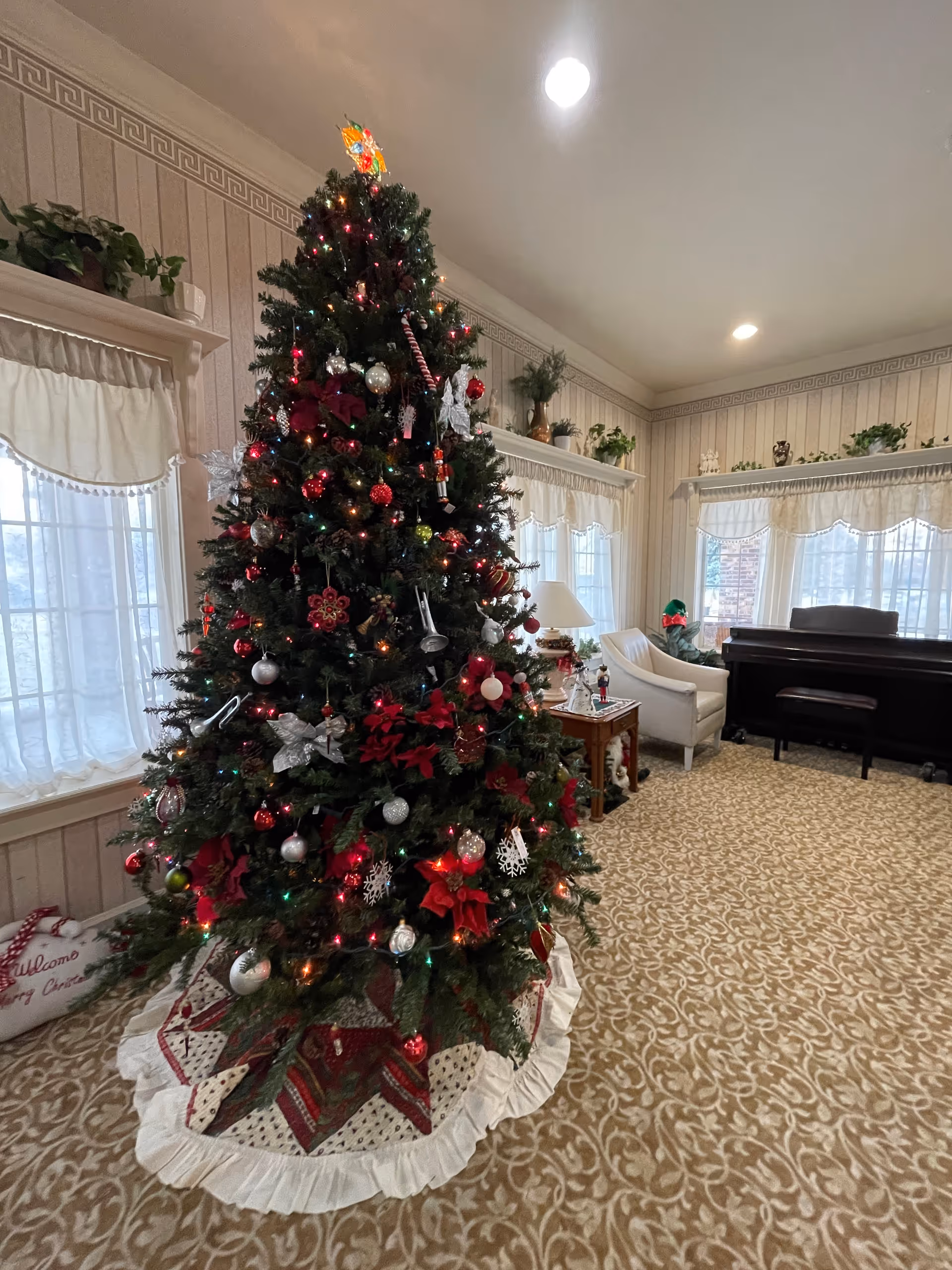 A decorated Christmas tree with red, silver, and white ornaments and multicolored lights stands in a carpeted room with patterned wallpaper. The room has large windows with white curtains, a white armchair, a wooden side table with a lamp, and a dark piano with a bench. There are plants on shelves above the windows and a festive holiday pillow on the floor near the tree.