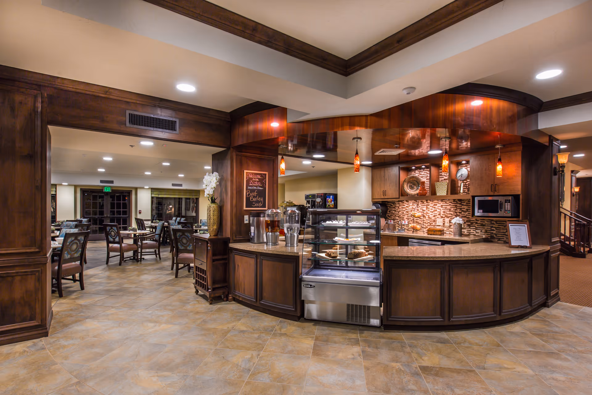 Interior view of a dining area and a small bistro counter with a glass display case containing pastries. The space features warm wooden cabinetry, pendant lighting, tiled flooring, and several tables and chairs arranged for dining.
