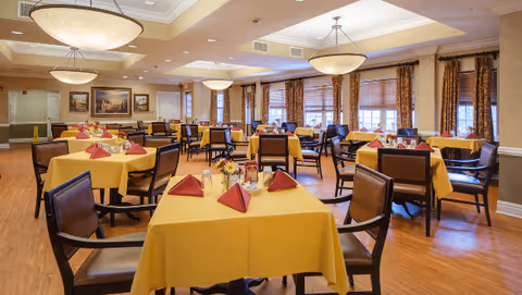 Bright dining room with multiple tables covered in yellow tablecloths and red folded napkins, surrounded by chairs under pendant lights.