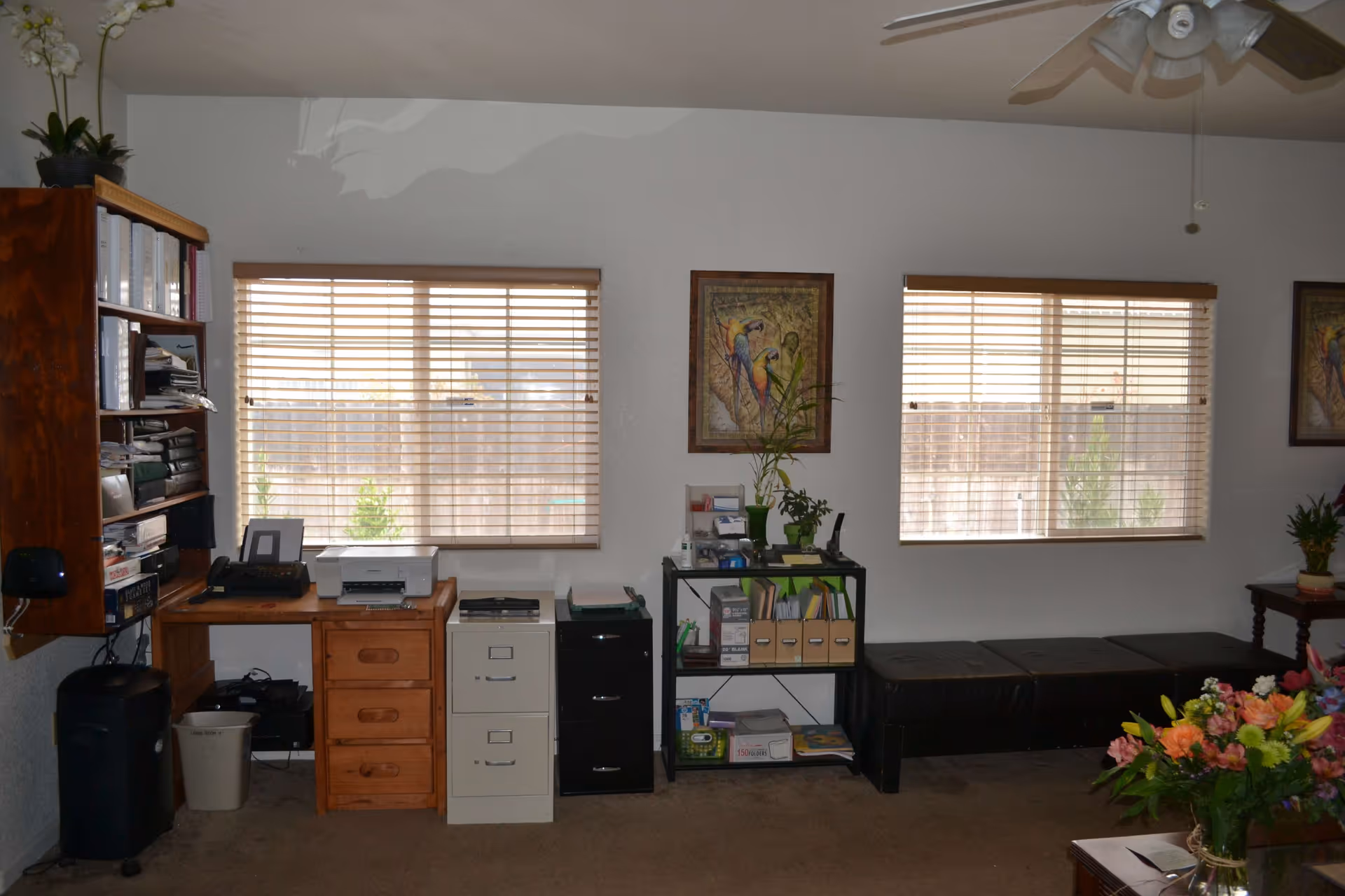 An office area inside a residential facility with two windows covered by wooden blinds. The room contains a wooden desk with office equipment including a printer and telephone, two filing cabinets, a black shelving unit with office supplies and plants, a black bench, and a ceiling fan. There are framed pictures of colorful birds on the wall and a bouquet of flowers on a table in the foreground.