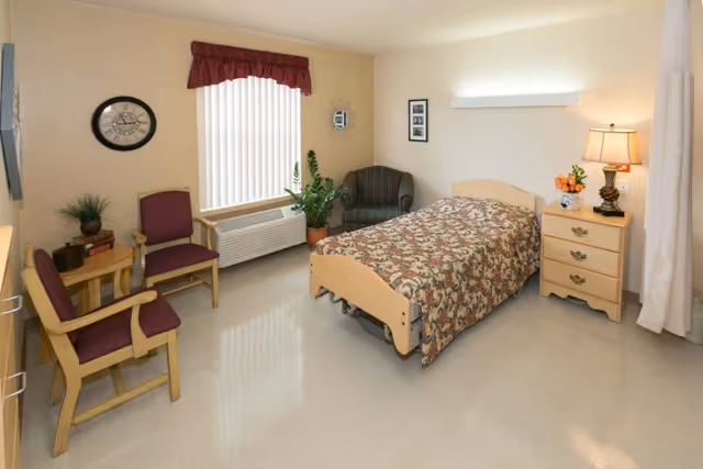 A simple, clean bedroom in a senior living facility featuring a single bed with a floral bedspread, a wooden nightstand with a lamp and flowers, two wooden chairs with purple cushions, a small round table with books and a plant, a striped armchair, a wall clock, and a window with vertical blinds and a red valance.