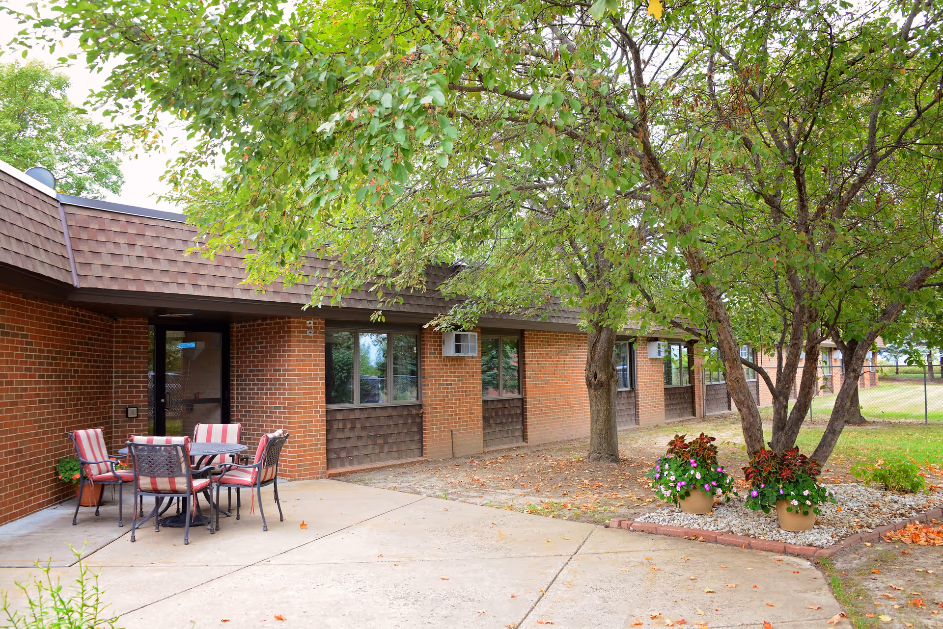 Outdoor patio area at Heritage Court with a round table and four chairs with red and white striped cushions. The patio is adjacent to a brick building with several windows and a door. There are trees providing shade and two flower pots with colorful flowers on a small landscaped area next to the patio.