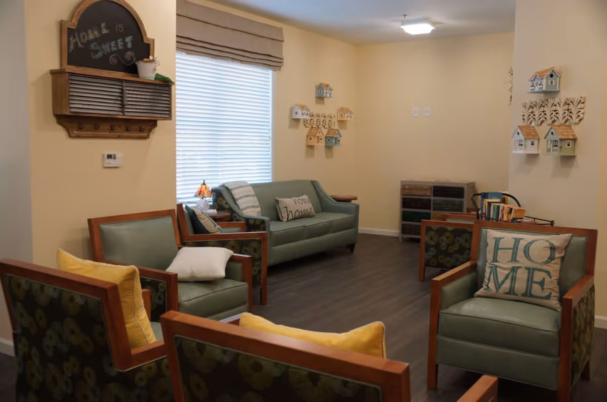 A cozy senior living facility common area with green cushioned chairs and a matching loveseat arranged around the room. The walls are decorated with small birdhouse wall hangings and a wooden wall organizer with a chalkboard that reads 'Home is Sweet'. There are pillows on the chairs, one with the word 'HOME' and another with the word 'home'. A window with blinds and a beige roman shade lets in natural light.