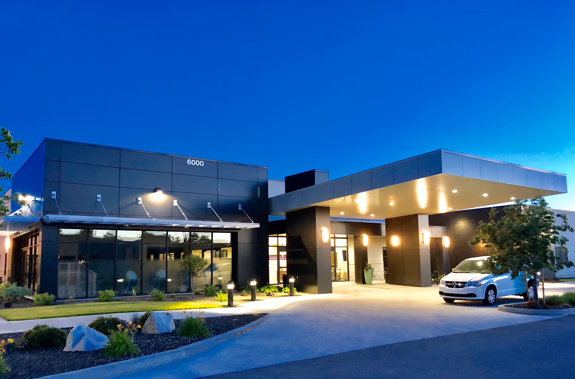 Exterior view of a modern senior living facility building at dusk with illuminated entrance canopy, large windows, landscaping with rocks and plants, and a white vehicle parked near the entrance.