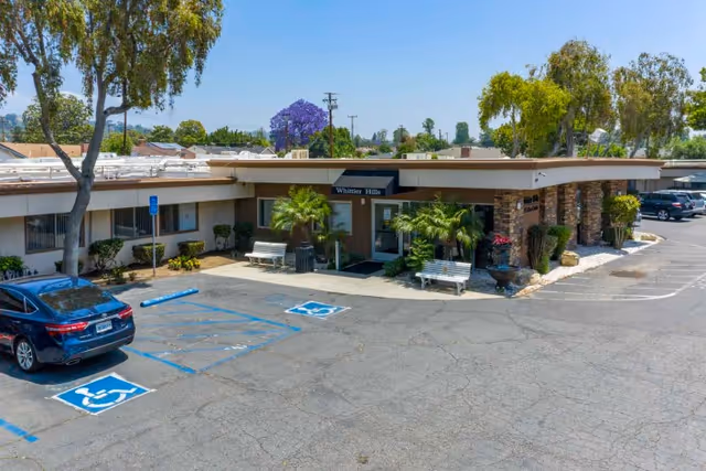 Exterior front entrance of Whittier Hills Healthcare Center showing the building facade, parking lot with handicap spaces, benches, and landscaping.