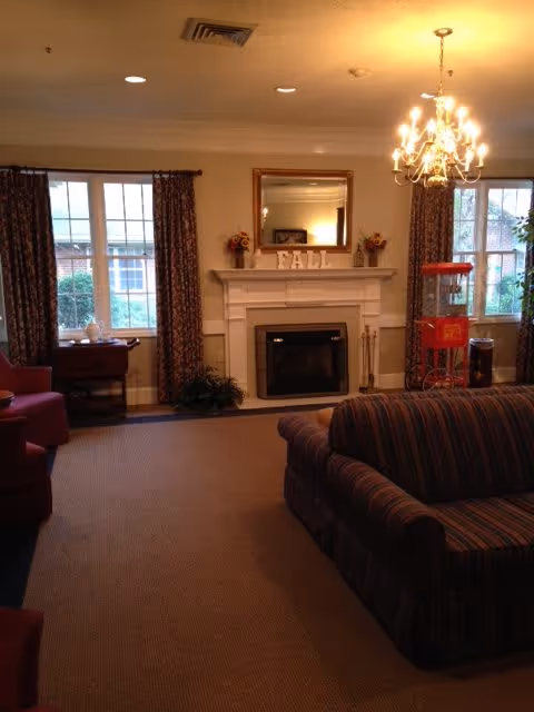 Cozy senior living community common area with a fireplace decorated with a mirror and fall-themed decor on the mantel. Two large windows with patterned curtains flank the fireplace. There is a striped couch in the foreground and a red popcorn machine near one window. A chandelier hangs from the ceiling providing warm lighting.