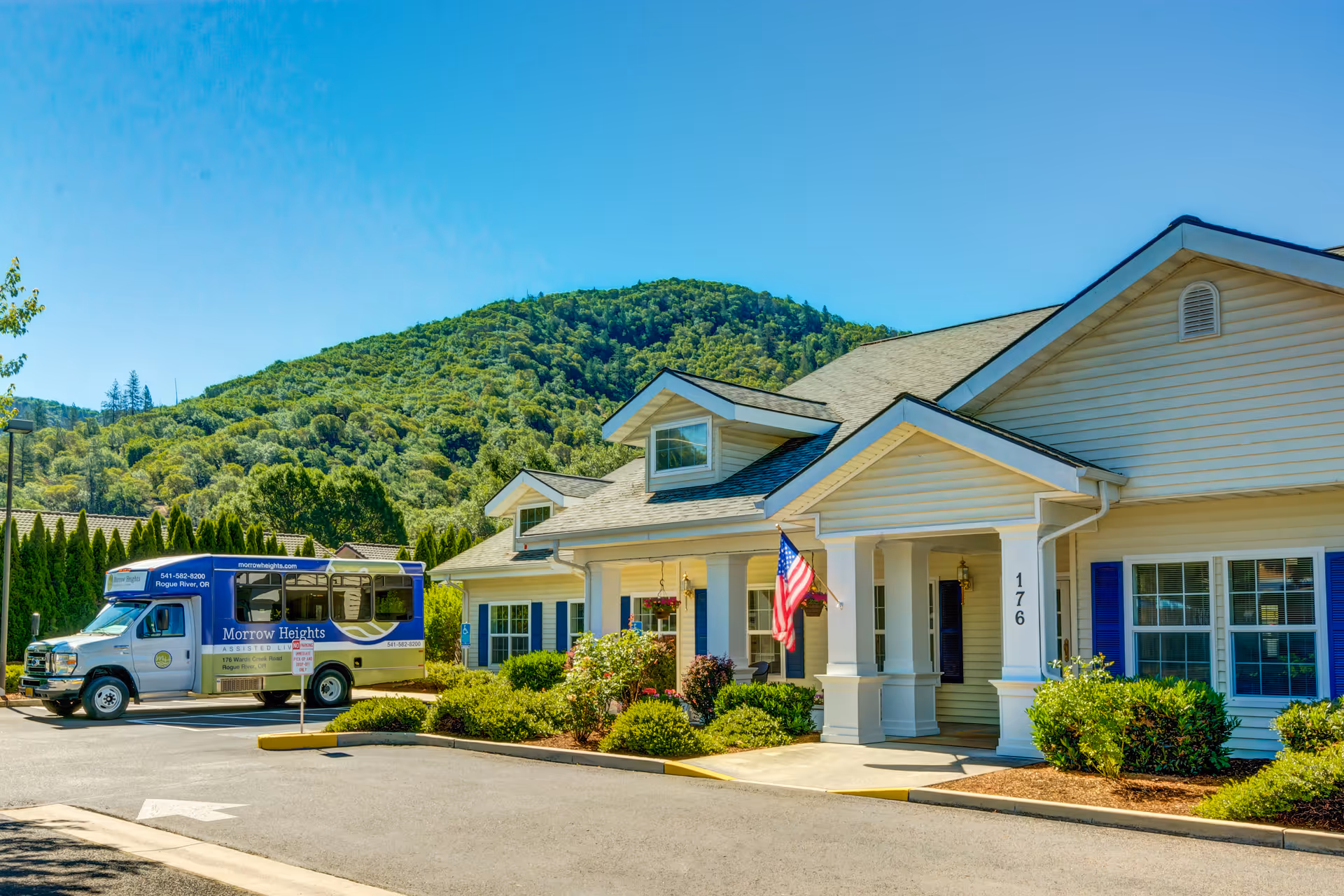 Exterior view of Morrow Heights Assisted Living facility with a beige building featuring blue shutters and a porch with an American flag. A blue and white shuttle bus is parked in front, and green hills are visible in the background under a clear blue sky.