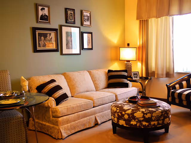 A warm living room with a beige sofa, striped throw pillows, a floral round ottoman, side table with a lamp, and framed pictures on the wall.