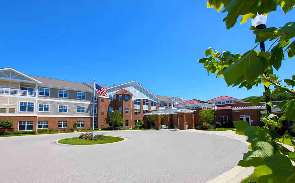 Exterior view of Wittenberg Village senior living facility on a clear sunny day, showing a large multi-story building with brick and beige siding, multiple windows, an American flag on a flagpole, a circular driveway, and green landscaping with trees and bushes.