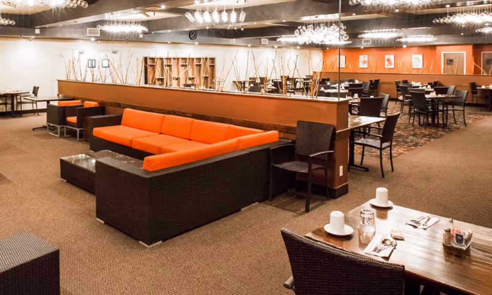 Interior view of a senior living facility dining and lounge area with a large orange sectional sofa, dark wicker chairs, wooden tables set with cups and utensils, decorative sticks in planters, and modern ceiling lights.