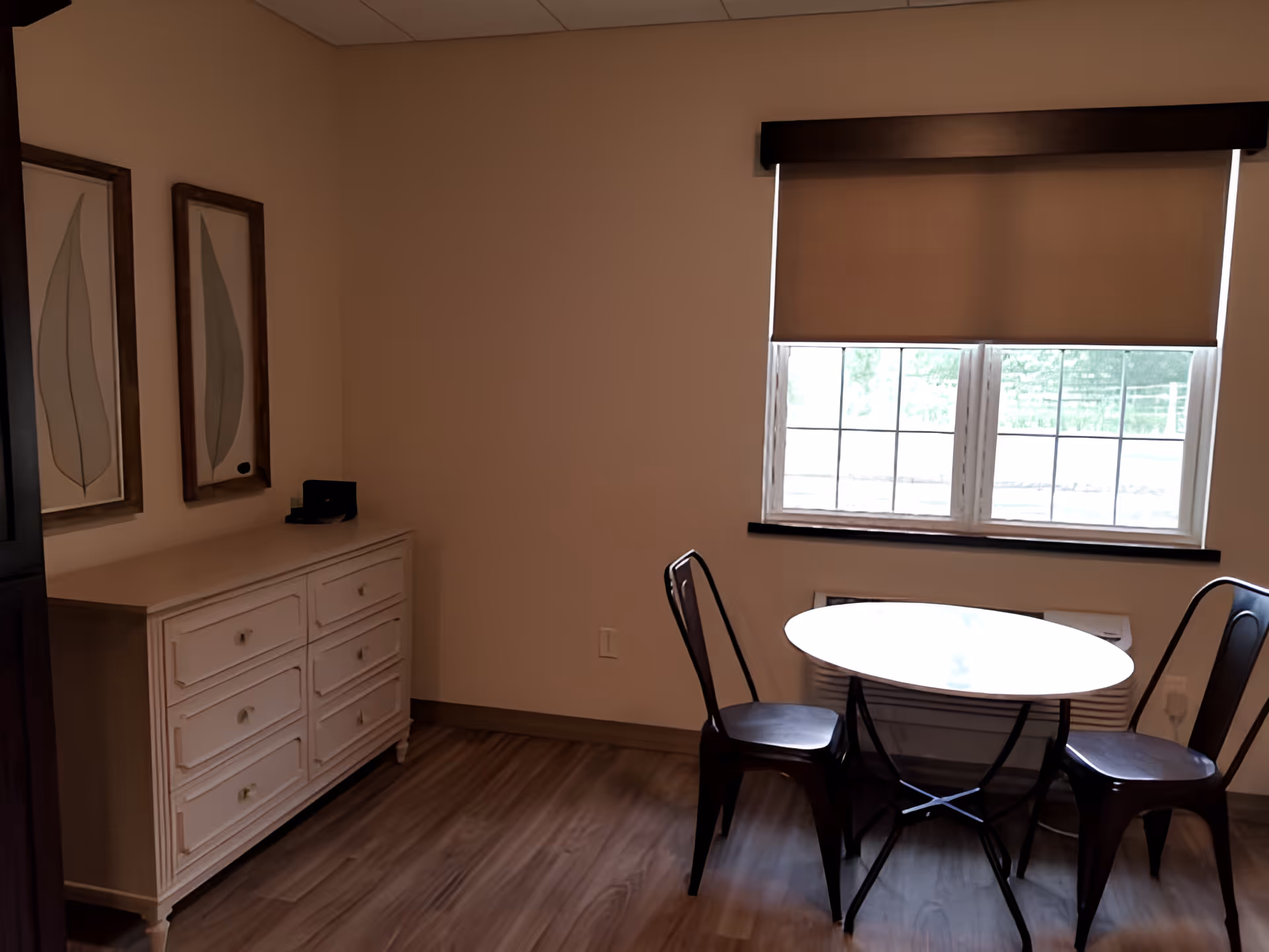 Small interior room with a round table and two chairs by a window, a white dresser and framed artwork on the wall.
