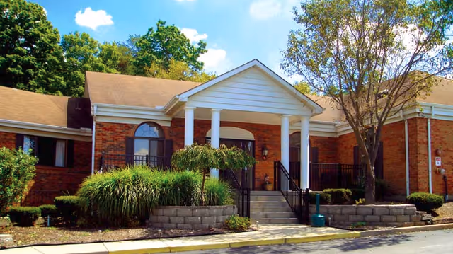 Front entrance of a brick single-story building with a white columned portico, steps, shrubs, and trees under a blue sky.