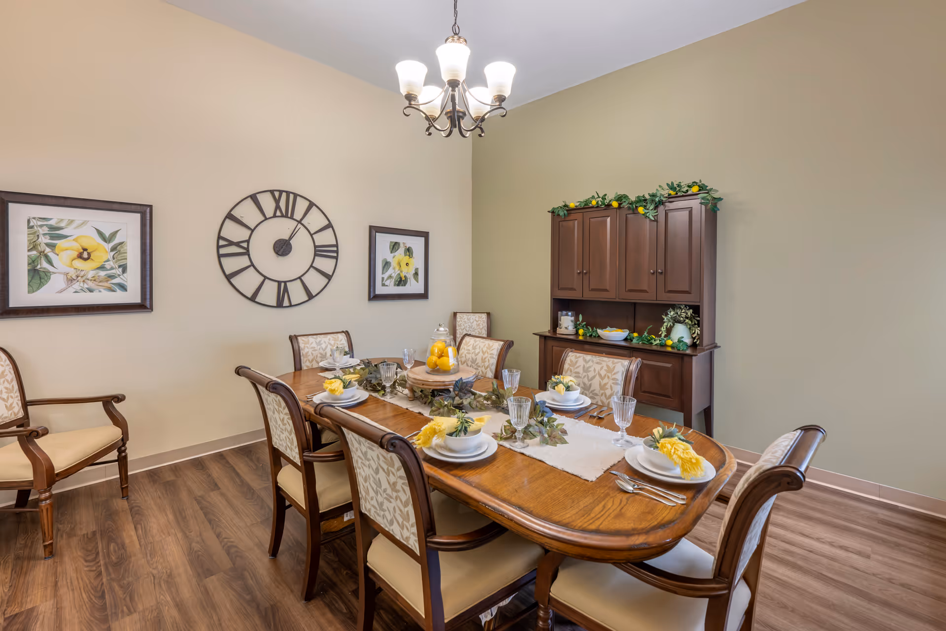 A dining room with a wooden dining table set for six people. The table is decorated with yellow flowers, greenery, and a centerpiece of lemons in a glass jar. There are six upholstered chairs around the table. On the wall behind the table, there is a large round clock and two framed floral paintings. A wooden cabinet with decorative greenery and lemons on top is against the wall. The room has wood flooring and a chandelier with five lights hanging from the ceiling.