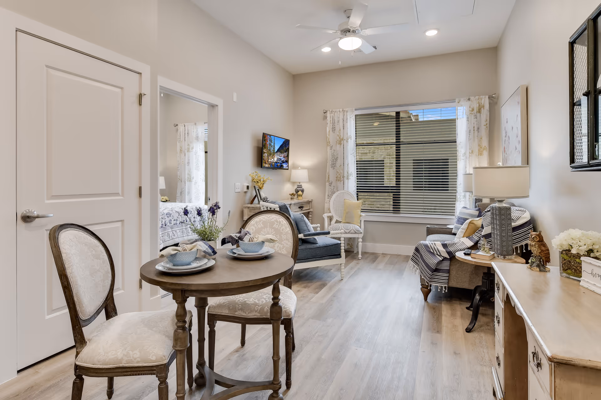 A cozy living area in a senior living facility with a small round wooden table set for two with bowls and napkins, two upholstered chairs, a blue armchair, a white chair with a yellow pillow, a gray sofa with a striped throw blanket, a wooden sideboard with a lamp and decorative items, a wall-mounted TV, and a large window with floral curtains letting in natural light.