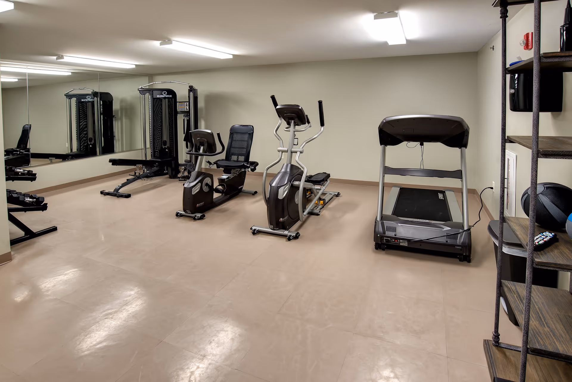 A small fitness room with exercise equipment including a treadmill, a recumbent bike, an elliptical machine, and a weight machine. The room has beige walls, a large mirror on one side, and a shelving unit with a medicine ball and remote controls.