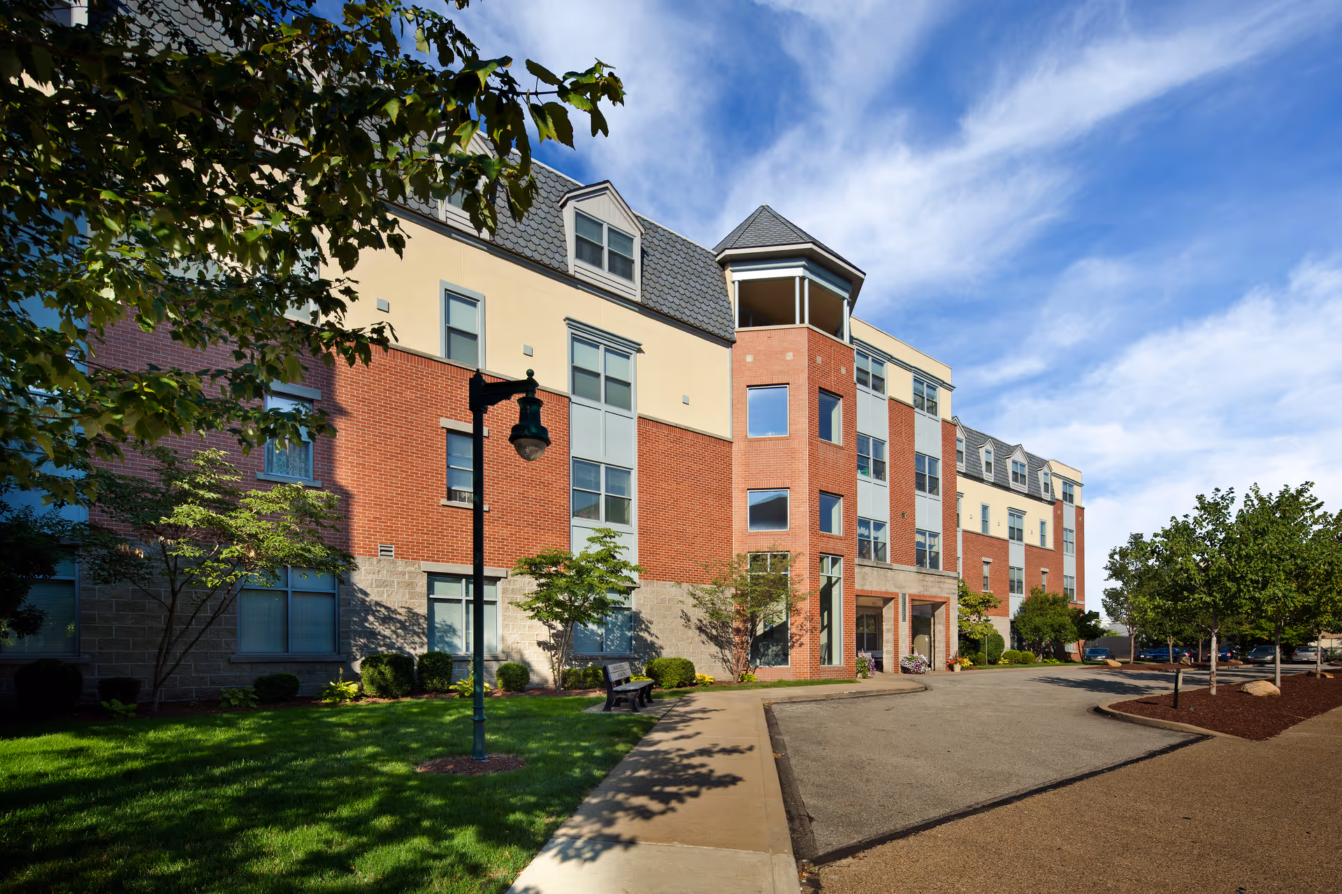 Three-story brick and stucco residential building with a landscaped lawn, walkway, benches and trees under a blue sky.