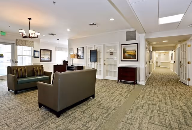 A spacious and well-lit common area in a senior living facility featuring two sofas with striped cushions, a carpeted floor, framed artwork on the walls, a lamp on a small table, and a hallway leading to other rooms.