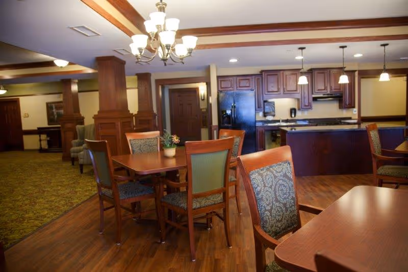 Interior view of a senior living facility dining area with wooden tables and chairs, a kitchen area with dark wood cabinets, a black refrigerator, and pendant lights hanging from the ceiling. The room has wooden flooring and a chandelier, with a carpeted area and additional seating visible in the background.